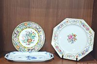 Three porcelain dishes displayed on wood shelf including octagonal floral platter, round floral bird plate, and rectangular blue sandwich tray