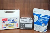 Photo shows the First Aid Kit, 50-piece package of black masks, and package of KN95 masks arranged side by side against a wood background.