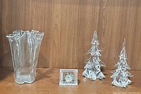 Clear view of all four glass items arranged side by side on a wooden shelf: ribbons vase, glass clock, pair of glass trees