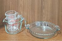 Photo showing 3 Pyrex glass measuring cups nested inside each other standing beside a round Pyrex glass pie plate with handles.