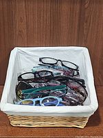 Multiple assorted used glasses displayed in a white linen-lined wicker basket showing a variety of frame colors and styles.