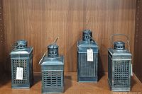 Four metal lanterns displayed on wood surface, includes three square and one taller rectangular lanterns all with handles and vented tops, showing overall lot content.