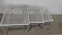 Three white metal patio nesting tables with diamond mesh tops arranged nested, showing the largest, medium, and smallest tables from an angled top side view.