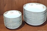 Stack of 11 smaller dessert bowls and 12 larger soup bowls with floral and gold trim pattern on the rim, placed on wood surface.
