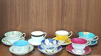 Seven porcelain teacups and saucers with a variety of colorful patterns and gold trim arranged on a wooden surface against a wooden wall.