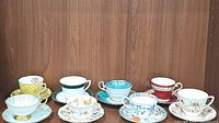 8 assorted porcelain teacups displayed side by side with matching saucers on wooden shelf showing variety of colors and designs.