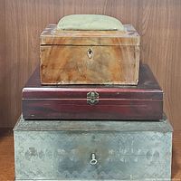 Three stacked vintage boxes: pin cushion sewing box on top, cherry wood box in middle, silver metal box at bottom