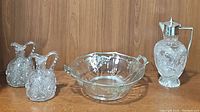 Group shot showing the pair of cut crystal cruets, the etched glass silver handled dish, and the cut glass decanter with silver top and handle on a wood shelf.