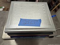 Stack of six coffee table books, top one with white/beige cover showing embossed design and blue tape labeled '25CP'.