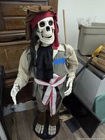 Full life sized pirate skeleton figure dressed in pirate hat, red head wrap, beige shirt, green vest, black cravat, brown pants and skeletal feet shoe covers, standing upright on a round black base.