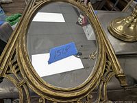 Close-up of oval mirror with bronze-colored ornate frame and blue tape showing '1528P' on mirror surface
