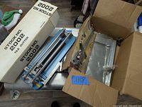 Full view of tile cutter in original box alongside a cardboard box with various metal parts and tools inside