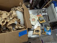 Wide view of the lot showing a cardboard box with frosted glass light shades wrapped in paper, two small floral trays, two refrigerator thermometers (one in original packaging), a wooden-handled hand saw, and a metal cabinet latch on a table.