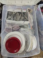 Top down view showing boxed stainless steel utensil set, red and white plastic plates, and cups inside a plastic tote.