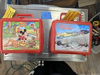 Photo showing two vintage lunch boxes side by side on a silver surface: one with Disney City Zoo design and one with racing car design, both with red handles and tags.