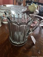 Photo of vintage clear pattern glass water pitcher showing scalloped vertical panels, thick glass, and curved handle with pouring spout.