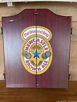Front view of the closed dart board cabinet showing Newcastle Brown Ale logo and branding on double doors with visible wood grain and side handles.
