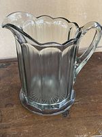 Clear glass pitcher with scalloped edge and vertical panel pattern seen from side on wooden surface.