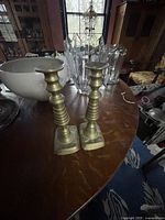 Pair of brass candlesticks on a wooden table with some decorative glass and ceramic items in background
