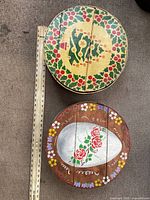 Top view of two round wooden cheese baskets side by side showing their hand painted holiday designs and measuring tape for scale