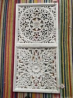 Two stacked white hand carved wooden panels with intricate floral and circular patterns shown against a striped fabric background.
