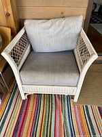 Front view of beige painted wicker arm chair with beige cushioned upholstery and woven wicker sides.
