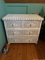 Front view of the vintage white wicker four-drawer chest showing woven wicker drawer fronts with round knob handles and wood flooring background.