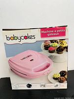Front view of the boxed pink Babycakes cupcake maker showing the appliance, cupcakes, and branding