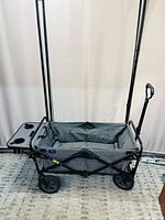Foldable wagon with fabric body, black metal frame, and four wheels. Tray attached at one side with cup holders. Handle for pulling visible.