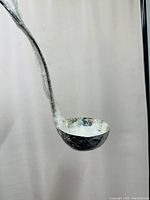 Photo of a silver skimming ladle with a long curved handle ending in a round bowl, displayed hanging against a light background.