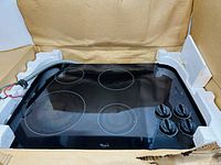 Top view of Whirlpool glass electric cooktop with four burners and attached wiring inside protective packaging.