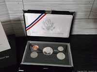 Opened black hinged box with white padded lid and red, white, and blue ribbon containing coins in clear display case with black background and silver lining; coins include copper penny and four silver coins.