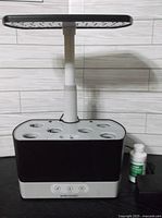 Front view of AeroGarden indoor hydroponic garden with adjustable light panel and seven plant slots, including a bottle of plant food beside it.