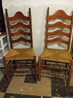 Two antique wooden ladder back chairs side by side with rush seats and turned wood spindles. Visible wear on wood finish.