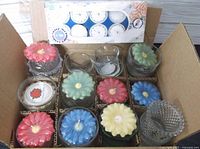 Box open showing 9 flower shaped candles in various colors, assorted glass candle jars, and box of tea lights