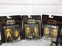 Three unopened double packs of commemorative gold Star Wars action figures laid side by side showing overall product packaging and contents.