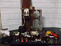 Photo showing a black table with wooden vintage-style jewelry box holding various necklaces and bracelets, earrings in small boxes and on display, plus colorful bangles and pendant necklaces arranged around it.