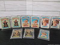 Front view of all nine Garbage Pail Kids cards in individual plastic protective cases, shown upright on a wooden surface.