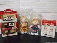 Photo showing all lot items: three diecast vehicles in original packaging, two soft dolls, and boxed Campbell Kids salt and pepper shaker set.