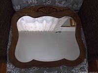 Front view of a vintage wall mirror with a wooden frame showing carved shell detail and curved edges, reflecting a window with sheer curtains.