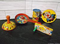 Five colorful vintage tin noise makers arranged on a black surface, showing clowns and balloons lithographs