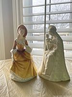 Both porcelain figurines displayed side by side, showing their size and detail under natural light by window blinds.
