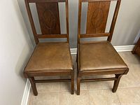Two wooden antique chairs with carved oval leaf detailing on the backrests and brown leather look padded seats, placed on tile floor near wall.