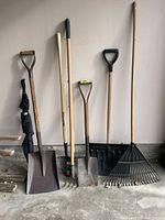 Multiple garden tools arranged upright against a wall on a concrete floor. Tools include shovels, forks, a rake, and a snow shovel.