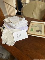 Lot contents showing folded assorted linen tablecloths and napkins stacked next to a framed print with a handwritten lot number 61 card.