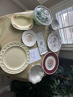 Overall image showing the collection of assorted plates and bowls arranged on a round table with a beige tablecloth. Variety of decorative ceramic and porcelain items with floral and textured designs.