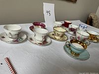 Front view of 11 assorted fine china tea cups with matching saucers arranged in a row on white tablecloth, showing floral and gold decorative patterns.