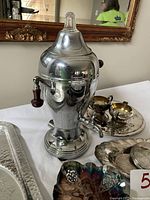 Close-up of Farberware electric coffee percolator with wooden handles and glass lid knob. Adjacent small silver plated sugar and creamer set and oval silver trays visible.