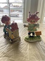 Photo showing two small Goebel ceramic statues, one of a red-headed child holding daisies with a Keep Off sign and another of a boy pushing a dog on a wooden cart.