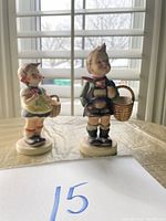 Photo of two West Germany Hummel porcelain figurines on a table in front of a window showing a sister and village boy each holding small baskets.